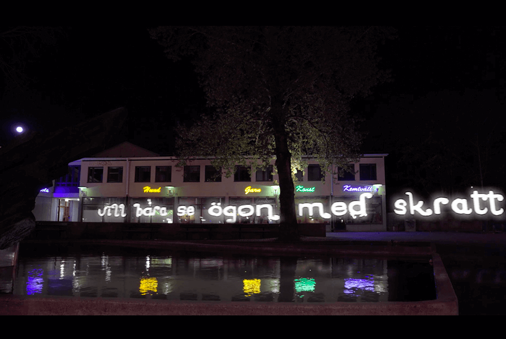 "Vill bara se ögon med skratt i" skrivet med ljus i mörkret med Bandhagens centrumhus i bakgrunden